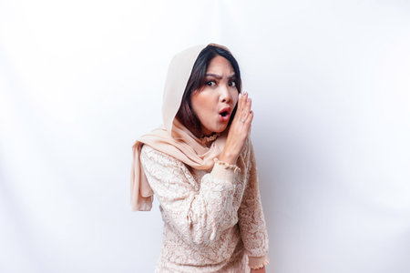 Young beautiful Asian Muslim woman wearing a hijab shouting and screaming loud with a hand on her...