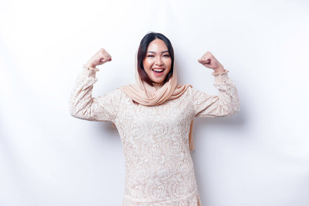 Excited Asian Muslim woman wearing a hijab showing strong gesture by lifting her arms and muscles smiling proudlyの写真素材