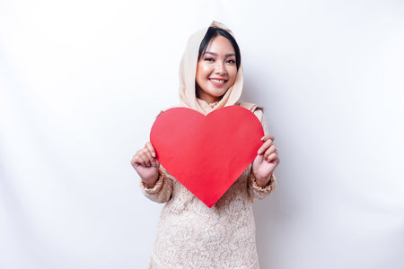 A happy young Asian Muslim woman wearing a hijab feels romantic shapes heart gesture expressing tender feelings and holding a red heart-shaped paperの写真素材