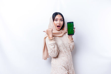 Shocked Asian woman wearing hijab, showing copy space on her phone screen, isolated by white backgroundの写真素材