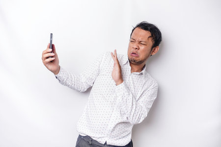 Young Asian man isolated on white background, looks depressed, face covered by arms frightened and nervous while holding his phoneの写真素材