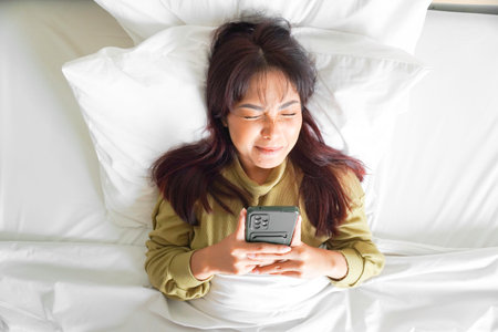 A thoughtful young Asian woman is holding her phone while lying on the bedの写真素材