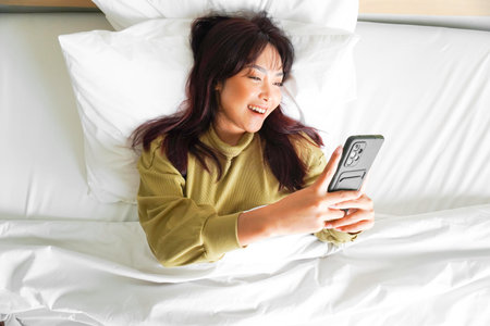 A smiling young Asian woman is laughing while holding her phone and lying on the bedの写真素材