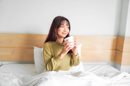 Image of beautiful young Asian woman wearing sweatshirt smiling and drinking tea on bed in white apartmentの写真素材