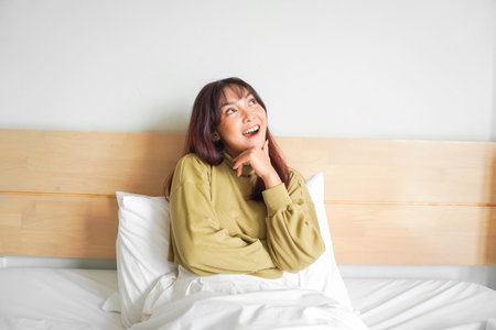 A thoughtful young Asian woman is looking aside and holding her chin while sitting on the bed