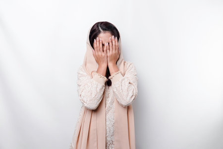 Young Asian Muslim woman isolated on white background, looks depressed, face covered by arms frightened and nervous.の写真素材