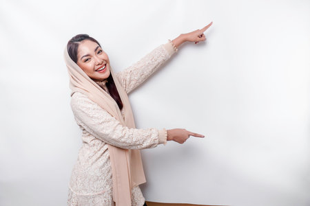 Young Asian Muslim woman smiling while pointing to copy space beside her