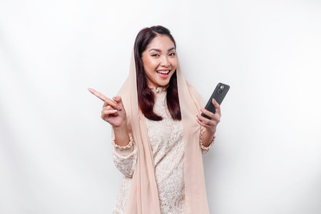 Excited Asian woman wearing hijab pointing at the copy space beside her while holding her phone, isolated by white backgroundの写真素材