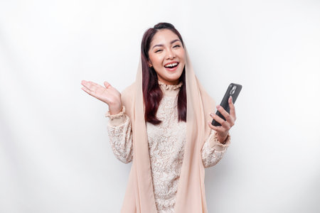 Excited Asian woman wearing hijab pointing at the copy space beside her while holding her phone, isolated by white backgroundの写真素材