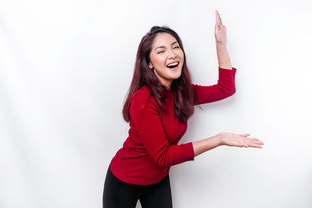 Excited Asian woman dressed in red, pointing at the copy space beside her, isolated by white backgroundの写真素材