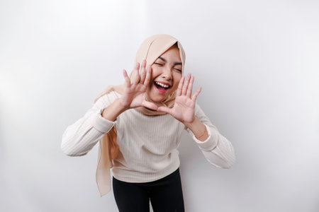 Young beautiful Asian Muslim woman wearing a headscarf shouting and screaming loud with a hand on her mouth. communication concept.の写真素材