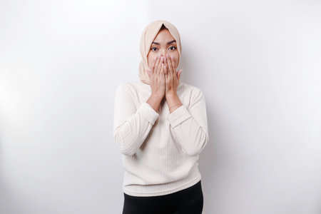 Portrait of a young Asian Muslim woman wearing a headscarf is tell be quiet, shushing with serious face, hush with finger pressed to lips, isolated by white backgroundの写真素材
