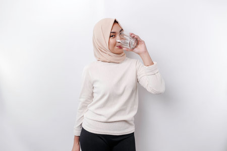 Joyful Asian Muslim woman wearing headscarf is drinking a glass of water, isolated on white background.の写真素材
