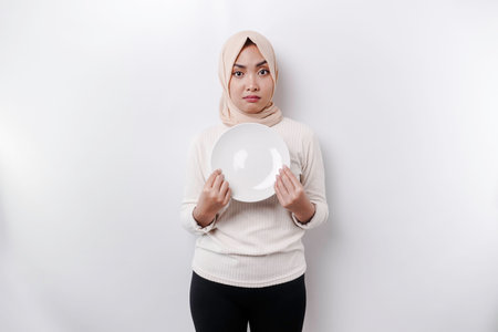 An Asian Muslim woman is fasting and hungry and holding a plate while looking aside thinking about what to eatの写真素材