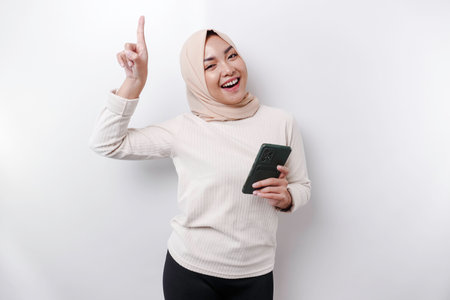 Excited Asian woman wearing hijab pointing at the copy space on top of her while holding her phone, isolated by white backgroundの写真素材