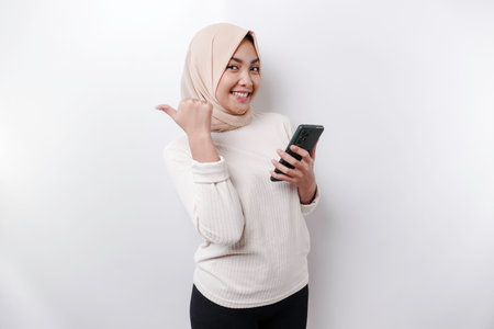 Excited Asian woman wearing hijab pointing at the copy space beside her while holding her phone, isolated by white backgroundの写真素材