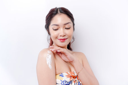 Skin Care Products Concept. Asian woman applying moisturizing lotion on body after shower, standing wrapped in towel, cropped imageの写真素材