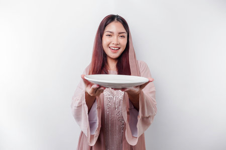 A smiling Asian Muslim woman is fasting and hungry and holding and pointing to a plateの写真素材