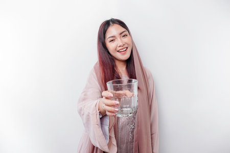 Joyful Asian Muslim woman wearing headscarf is drinking a glass of water, isolated on white background.の写真素材