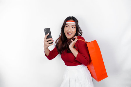 An attractive Asian woman standing excited holding an online shopping bag and her smartphone, studio...