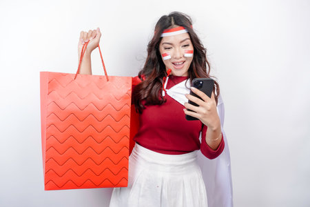 An attractive Asian woman standing excited holding an online shopping bag and her smartphone, studio shot isolated on white background. Indonesia's Independence day conceptの写真素材