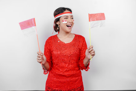 Happy smiling Indonesian woman wearing red kebaya and headband holding Indonesia's flag to celebrate...