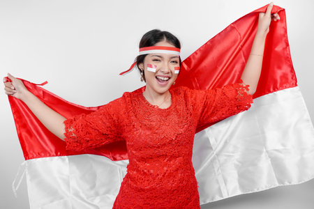 Happy smiling Indonesian woman wearing red kebaya and headband holding Indonesia's flag to celebrate Indonesia Independence Day isolated over white background.の写真素材