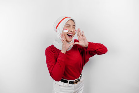 Young beautiful muslim woman wearing a red top and white hijab is shouting and screaming loud with a hand on her mouth. Indonesia's independence day concept.の写真素材
