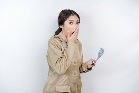 A shocked young government worker woman is wearing khaki uniform holding cash money in Indonesian rupiah isolated by white backgroundの写真素材