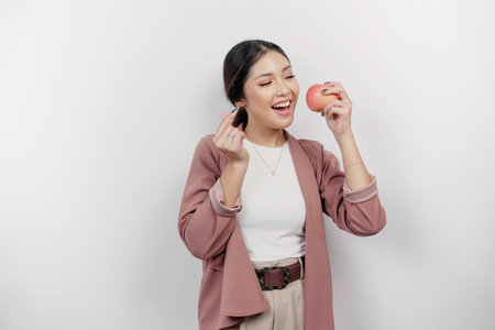 Lovely Asian woman employee is eating apple and gesturing love sign by her fingers, isolated by white background.の写真素材