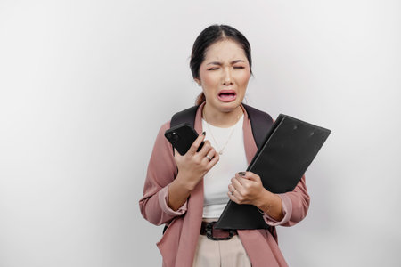 A stressed young Asian schoolgirl wearing cardigan while wearing a backpack and handphone, isolated by white background.の写真素材