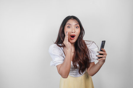 A young beautiful Asian woman is shouting and screaming loud with a hand on her mouth while holding her phone, isolated by white background.の写真素材