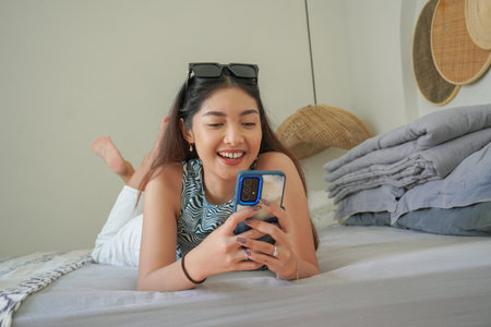 Smiling Asian woman laying down on bed with sunglasses on top while using mobile phone, exploring social media in the morningの写真素材