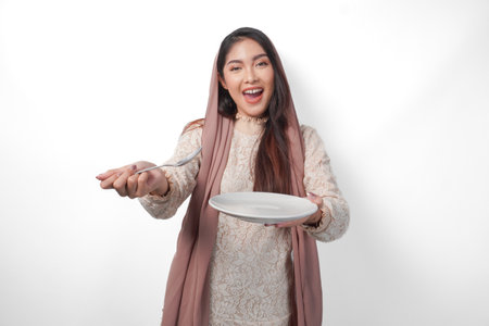 Excited Asian Muslim woman holding plate and cutlery to eat after fasting while smiling cheerfully on isolated white background. Ramadan conceptの写真素材