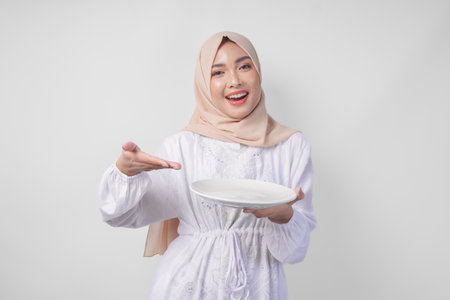 Smiling young Asian Muslim woman in a veil hijab pointing to an empty plate with copy space over it, presenting food menu for iftar. Ramadan conceptの写真素材