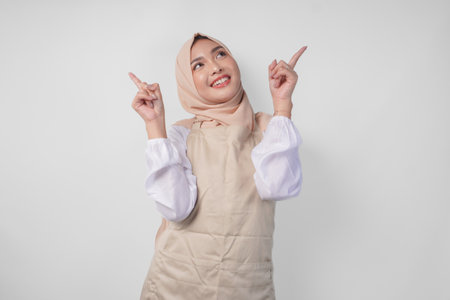 Overjoyed Asian woman wearing hijab and cream apron pointing up to the copy space above, standing over isolated white backgroundの写真素材