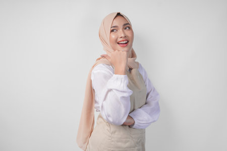 Excited young Asian Muslim woman in hijab and cream apron pointing to the empty copy space on the left and right side. Ramadan advertisement conceptの写真素材