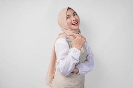 Excited young Asian Muslim woman in hijab and cream apron pointing to the empty copy space on the left and right side. Ramadan advertisement conceptの写真素材