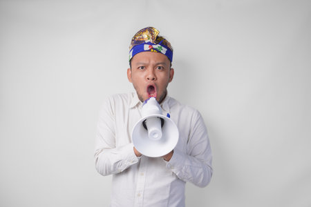 Overjoyed Balinese man in white shirt and traditional headdress shouting at megaphone feeling excited, isolated by white backgroundの写真素材