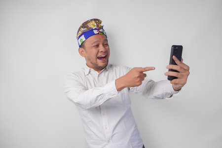 Young Balinese man in white shirt and traditional headdress doing video call with family or friend feeling overjoyed and excitedの写真素材