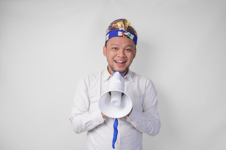 Energetic Balinese man in white shirt and traditional headdress shouting at megaphone while raising...
