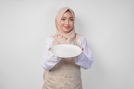 Beautiful young Asian Muslim woman in hijab and cream apron presenting an empty plate with copy space over it, showing the food menu for iftarの写真素材
