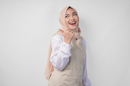 Excited young Asian Muslim woman in hijab and cream apron pointing to the empty copy space on the left and right side. Ramadan advertisement conceptの写真素材
