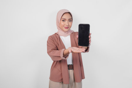 Young Asian Muslim woman wearing hijab is smiling while showing copy space on her smartphone screen, isolated by white background.の写真素材