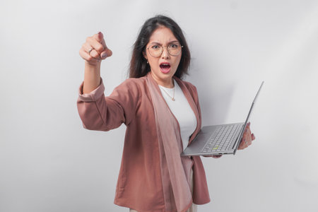 Enraged young Asian business woman holding a laptop pointing to the camera with angry expression, isolated by white background.の写真素材