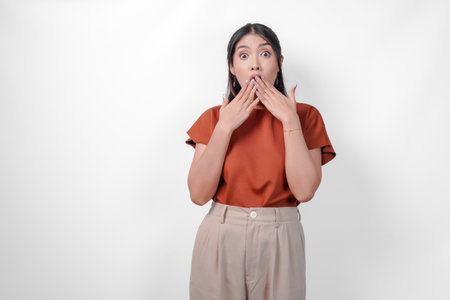 Surprised young Asian woman in a brown shirt and taupe pants is looking shocked with her hands covering her mouth, isolated by white background.の写真素材