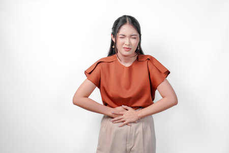 Unwell sick ill Asian woman in an brown shirt is holding her stomach. She looks uncomfortable and in pain. Concept of discomfort and unease.の写真素材