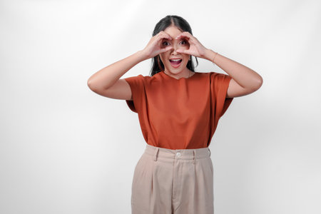 Happy young Asian woman wearing a brown shirt making binocular gesture with her hands over isolated white background.の写真素材