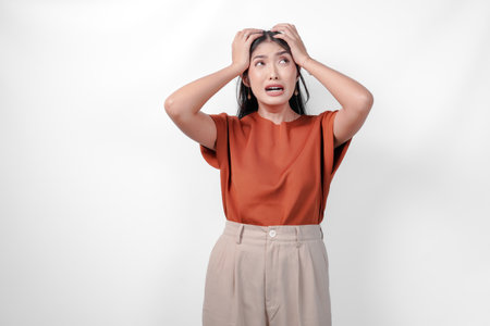 Stressed young Asian woman in a brown shirt and taupe pants is holding her head in her hands suffering from headache.の写真素材