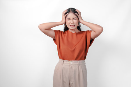Stressed young Asian woman in a brown shirt and taupe pants is holding her head in her hands suffering from headache.の写真素材
